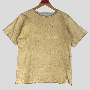 May include: Tan short-sleeve t-shirt with a speckled pattern and the text "STONE ISLAND" across the chest. The shirt is hanging on a wooden hanger.