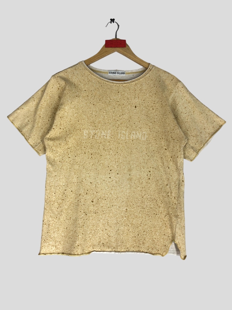 May include: Tan short-sleeve t-shirt with a speckled pattern and the text "STONE ISLAND" across the chest. The shirt is hanging on a wooden hanger.
