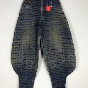 Pode incluir: Calças de ganga azuis com um design único e estampado. As calças têm cintura alta e uma forma de perna larga, tipo balão. Uma etiqueta vermelha pende da cintura. O denim tem um aspeto desbotado e desgastado.