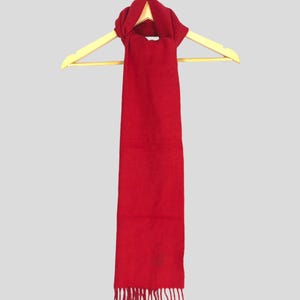 May include: A solid red scarf with a fringed bottom, hanging on a wooden hanger. The scarf is a deep shade of red and appears to be made of a soft material. The scarf is displayed against a light gray background.