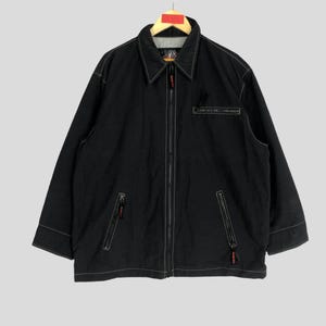 May include: Black jacket with white stitching and a full-length zipper. The jacket has a collared neckline, two front pockets, and a small logo on the chest. The jacket is hanging on a wooden hanger.