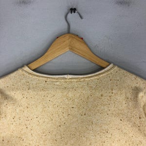 May include: A tan-colored sweatshirt with a speckled pattern, hanging on a wooden hanger. The sweatshirt has a white trim around the neckline. The background is a plain, light gray wall.