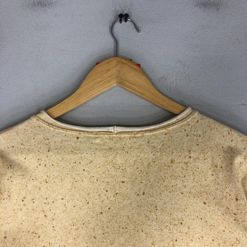 May include: A tan-colored sweatshirt with a speckled pattern, hanging on a wooden hanger. The sweatshirt has a white trim around the neckline. The background is a plain, light gray wall.
