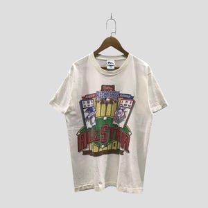 May include: A white short-sleeve t-shirt featuring a graphic print of the MLB All-Star Game. The design includes a baseball field illustration, the text "ALL STAR GAME," and scoreboards for the American and National leagues in red, blue, and green.