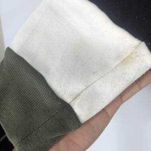 Może przedstawiać: Zbliżenie fragmentu rękawa bluzy. Rękaw ma blokowy design z białą g&oacute;rną częścią, czarnym g&oacute;rnym rogiem i oliwkowym mankietem. Tkanina wydaje się być miękkim materiałem dzianinowym.