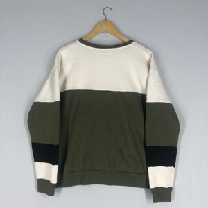 Może przedstawiać: Bluza w kolorowe bloki w kolorze kremowym, oliwkowym i czarnym. Top z długim rękawem wisi na drewnianym wieszaku na jasnoszarym tle. Bluza ma okrągły dekolt oraz prążkowany d&oacute;ł i mankiety.