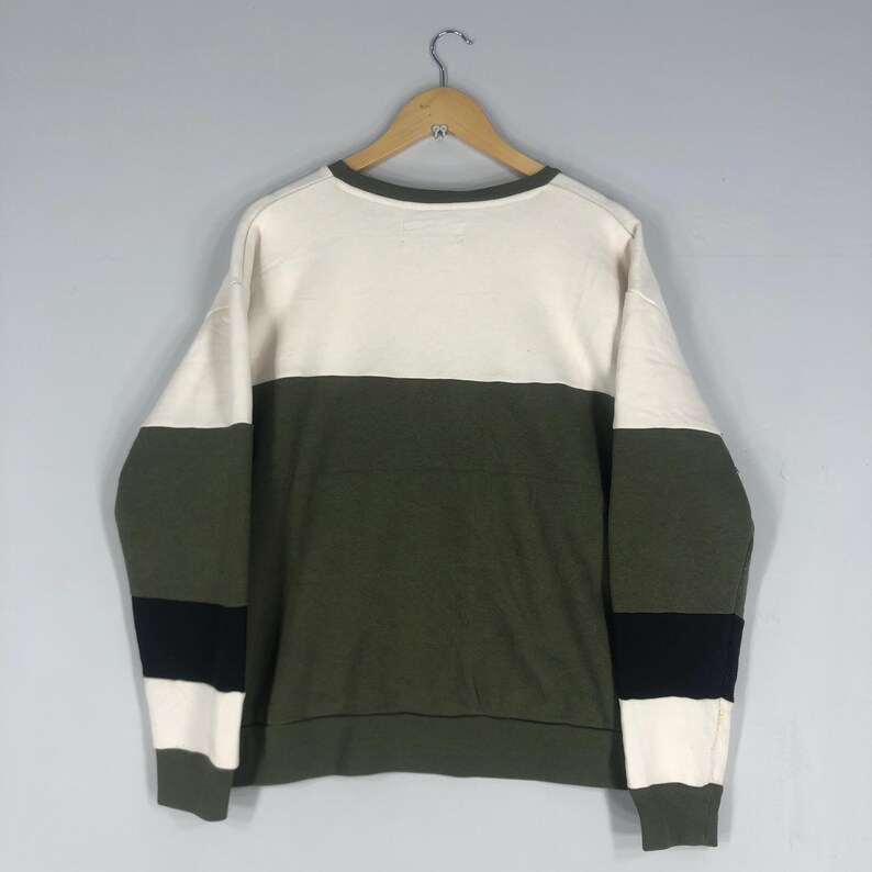 Może przedstawiać: Bluza w kolorowe bloki w kolorze kremowym, oliwkowym i czarnym. Top z długim rękawem wisi na drewnianym wieszaku na jasnoszarym tle. Bluza ma okrągły dekolt oraz prążkowany d&oacute;ł i mankiety.