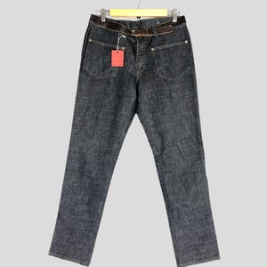 May include: Dark blue denim jeans with a brown leather belt and a red price tag hanging from the belt loop. The jeans are hanging on a wooden hanger.