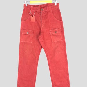 May include: Red denim pants with two front pockets and two cargo pockets, hanging on a hanger. The pants have a button closure and a small tag attached. The pants are a solid red color.