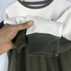 Może przedstawiać: Bluza w blokach kolor&oacute;w z białą g&oacute;rną częścią, ciemnozieloną dolną częścią oraz czarno-białym rękawem. Mankiety rękawa są ciemnozielone z białym akcentem.