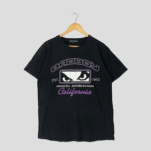 May include: Black t-shirt with a graphic design featuring the text "BADBOY" above an eye design, "EST. 1982", "LOS ANGELES", and "California" in purple. The shirt is hanging on a wooden hanger.