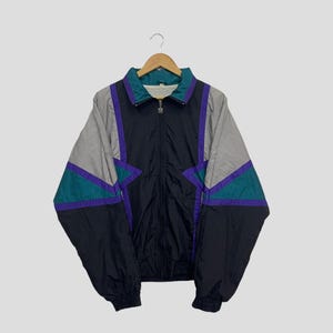 May include: A vintage black, gray, teal, and purple windbreaker jacket with a full zipper closure. The jacket features a color block design with geometric shapes and a teal collar. The jacket is hanging on a wooden hanger.