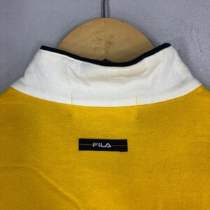 May include: Yellow polo shirt with a white collar and black trim. The shirt has a black Fila logo on the chest.