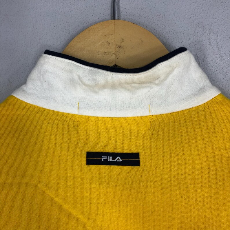 May include: Yellow polo shirt with a white collar and black trim. The shirt has a black Fila logo on the chest.