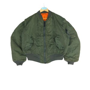 May include: A green bomber jacket with a zip closure and orange lining. The jacket has two pockets on the front and a zipped pocket on the left sleeve.