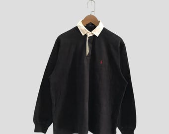 May include: A long-sleeve black rugby shirt featuring a contrasting white collar and a buttoned placket. The garment includes a small red embroidered logo on the left chest area and is displayed on a wooden hanger against a plain background.
