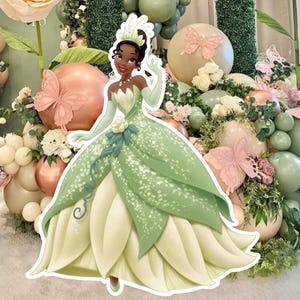 May include: A cartoon princess cutout in a green and white dress, tiara, and gloves is shown. The background features floral decorations, balloons, and large paper flowers in pink, green, and white, creating a whimsical party theme.