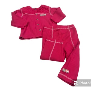 May include: A pink two-piece baby outfit with a long-sleeved cardigan and matching pants. The cardigan has a button closure and white stitching detail. The pants have white stitching detail and a small logo on the leg.
