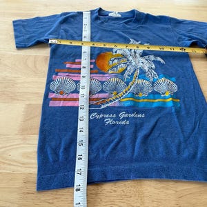 May include: A blue t-shirt with a graphic of a palm tree and seashells. The text "Cypress Gardens, Florida" is printed on the shirt.
