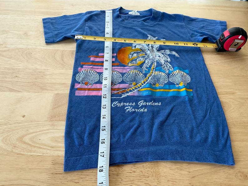 May include: A blue t-shirt with a graphic of a palm tree and seashells. The text "Cypress Gardens, Florida" is printed on the shirt.