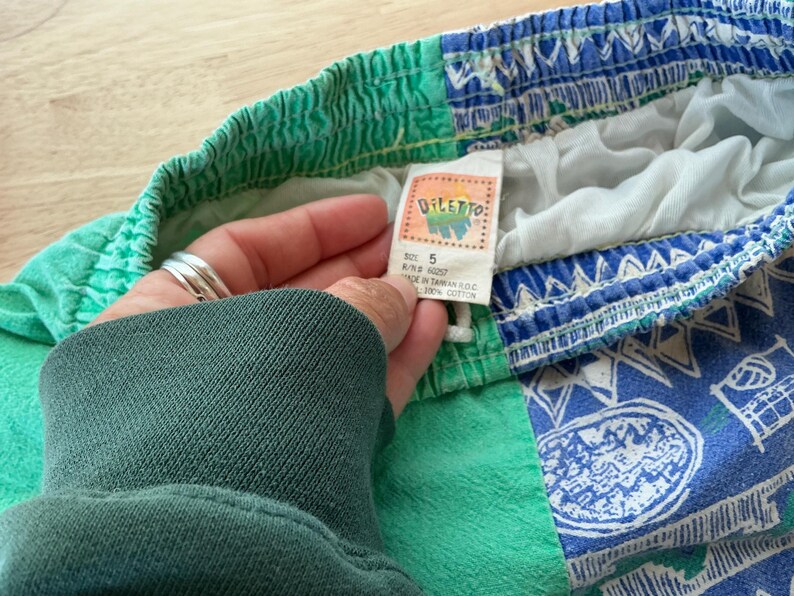 May include: Vintage children's shorts with a colorful design. The shorts feature a green waistband and a blue and white patterned lower section. The tag reads "DILETTO" and "SIZE 5". Made of 100% cotton.