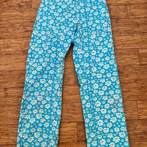 Può includere: Pantaloni blu con un motivo floreale a margherite bianche. I pantaloni hanno un taglio a gamba dritta e una chiusura con bottoni e cerniera. Le margherite hanno centri verdi.