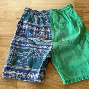 May include: Blue and green shorts with an elastic waistband. The left side features a blue pattern with white and green illustrations of palm trees, a creature, and a volleyball. The right side is solid green.