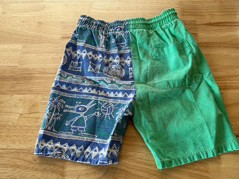 May include: Blue and green shorts with an elastic waistband. The left side features a blue pattern with white and green illustrations of palm trees, a creature, and a volleyball. The right side is solid green.