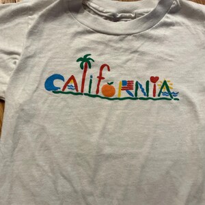 May include: White t-shirt with a colorful graphic that says "CALIFORNIA" with various symbols including a palm tree, an orange, a heart, and a wave.