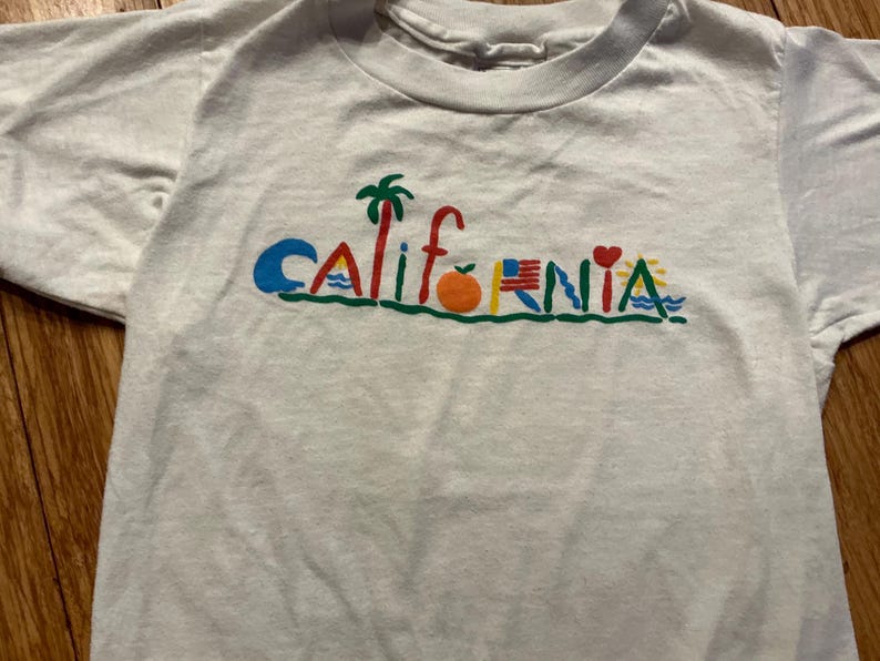 May include: White t-shirt with a colorful graphic that says "CALIFORNIA" with various symbols including a palm tree, an orange, a heart, and a wave.