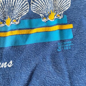May include: Blue t-shirt with a graphic of three seashells and yellow stripes. The text "Sunshine Souvenir 1988" is printed on the bottom right corner of the shirt.