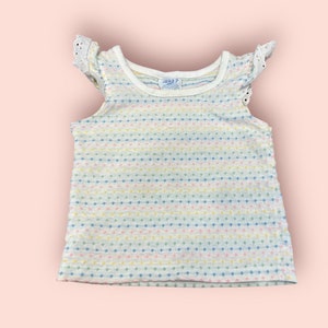 May include: A white sleeveless shirt with a rainbow polka dot pattern and white lace trim on the sleeves. The shirt has a round neckline and is made of a soft, stretchy fabric.