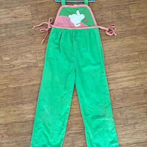 May include: Green and pink overall with a white bunny applique holding a carrot. The overall has adjustable straps and a tie at the waist.