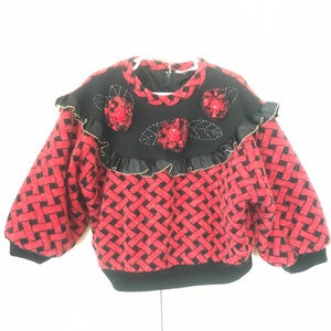 May include: A red and black patterned sweater with a black neckline and sleeves. The sweater features red floral embellishments and a ruffled trim with gold accents. The sleeves are puffy, and the sweater has a zipper closure.