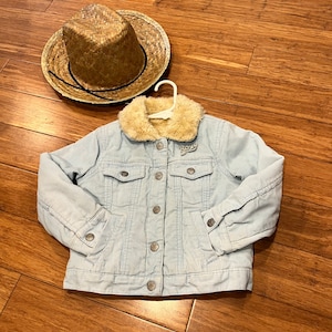 May include: A light blue corduroy jacket with a faux fur collar and silver buttons. The jacket has a striped pattern and is laid flat on a wooden surface. A straw cowboy hat is also in the image.