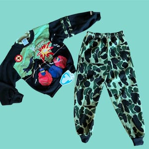 May include: Black sweatshirt with a graphic of boxing gloves and the word "VICTORY". The sleeves have a camouflage pattern. The matching pants are green and black camouflage. The set is displayed on a light blue background.