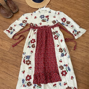 May include: A vintage-style white dress with a red floral pattern and a red center panel. The dress has long sleeves, a high collar, and a waist tie. A straw hat with a blue ribbon and brown cowboy boots are also in the image.