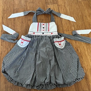 May include: A black and white gingham dress with red heart embroidery and white lace trim. The dress has a halter neckline and two white ribbon ties.