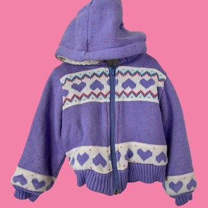 May include: A purple knitted zip-up jacket with a white and pink heart pattern. The jacket has a hood and a zipper closure.