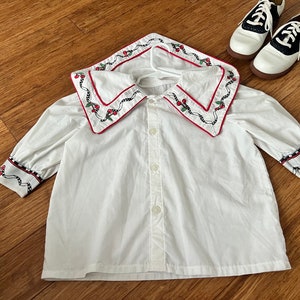 May include: White button-down shirt with a large, red-trimmed collar featuring a cherry and vine embroidery design. The shirt has long sleeves with a similar cherry and vine embroidery on the cuffs.
