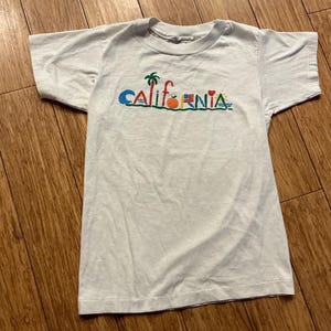 May include: White t-shirt with a colorful graphic of the word "CALIFORNIA" with various icons including a palm tree, an orange, a heart, and a surfboard.