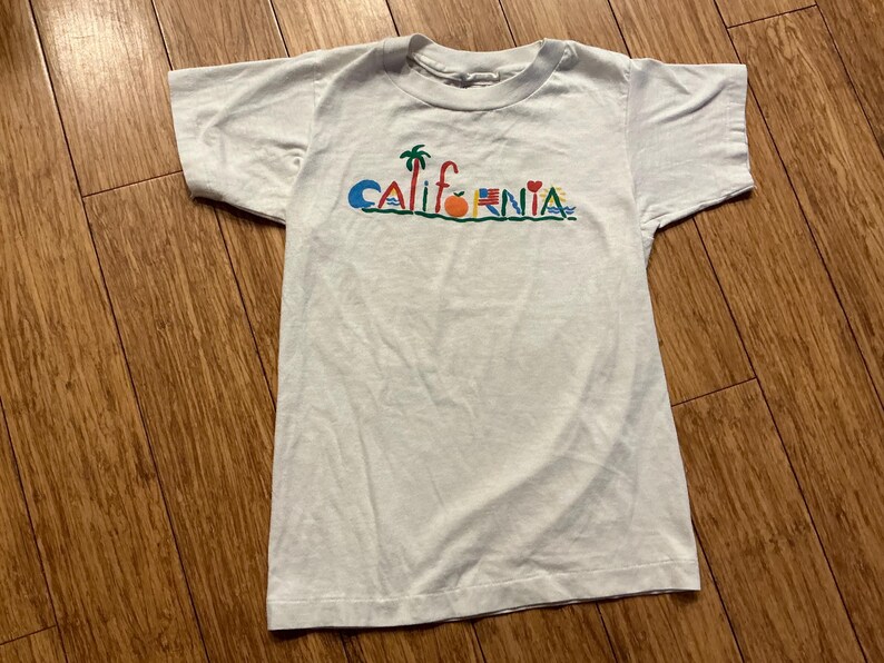 May include: White t-shirt with a colorful graphic of the word "CALIFORNIA" with various icons including a palm tree, an orange, a heart, and a surfboard.
