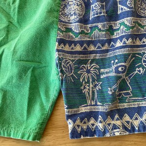 May include: Green and patterned shorts. The shorts are split, with one side solid green and the other featuring a blue and green print with palm trees, volleyballs, and a cartoon character. A pink measuring tape is visible.