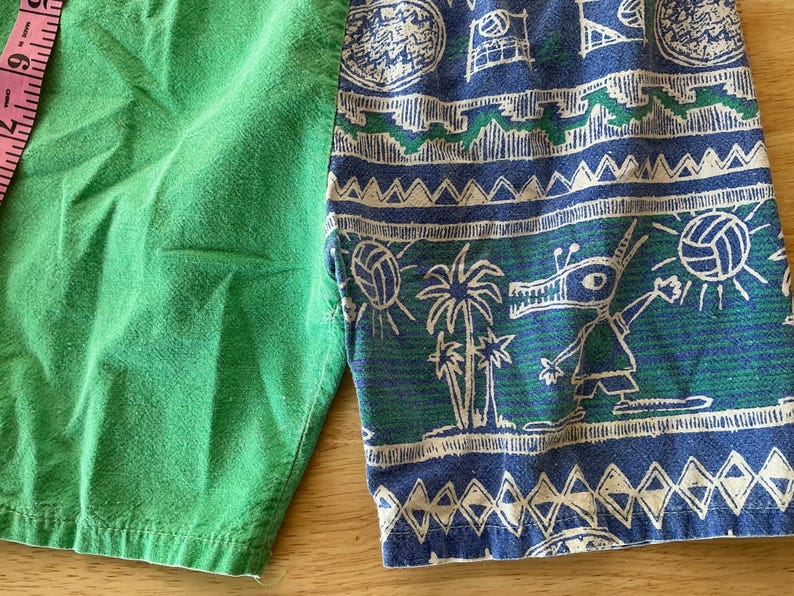 May include: Green and patterned shorts. The shorts are split, with one side solid green and the other featuring a blue and green print with palm trees, volleyballs, and a cartoon character. A pink measuring tape is visible.