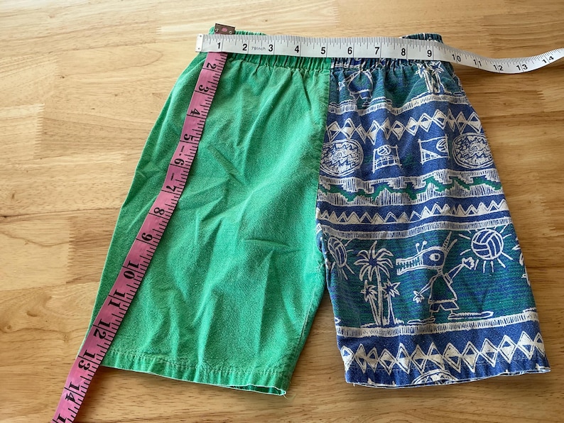 May include: Green and blue patterned shorts with a measuring tape. The shorts are divided into two sections: solid green and a blue pattern with white accents. The shorts measure approximately 14 inches across the waist.
