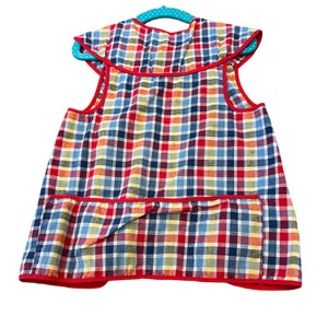 May include: A red, blue, and yellow plaid apron with a single pocket and red trim. The apron is hanging on a teal hanger.