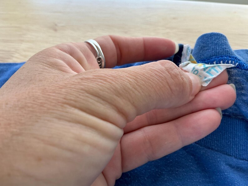 May include: Close-up of a blue t-shirt with a white tag attached to the collar. The tag has the words "100% Cotton" printed on it.