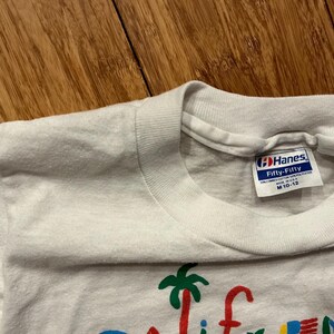 May include: White Hanes Fifty-Fifty t-shirt with a colorful graphic of a palm tree and the word "California". The tag says "Hanes Fifty-Fifty" and "M 10-12".