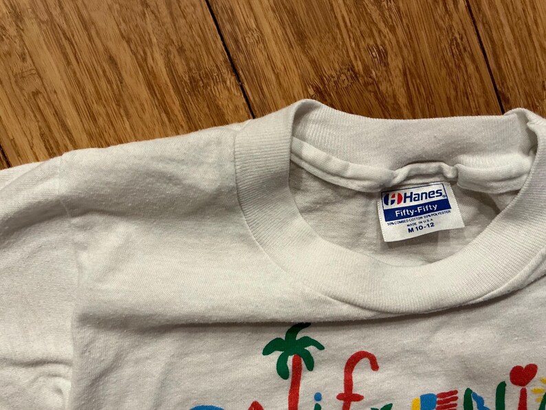 May include: White Hanes Fifty-Fifty t-shirt with a colorful graphic of a palm tree and the word "California". The tag says "Hanes Fifty-Fifty" and "M 10-12".