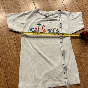 May include: White t-shirt with a colorful "California" graphic featuring palm trees, a surfboard, an orange, a flag, and a heart. The shirt is being measured with a tape measure.